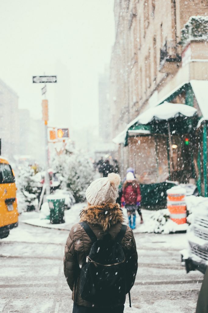 femme en ville sous la neige portant une doudoune un bonnet à pompon et une sac à dos noir